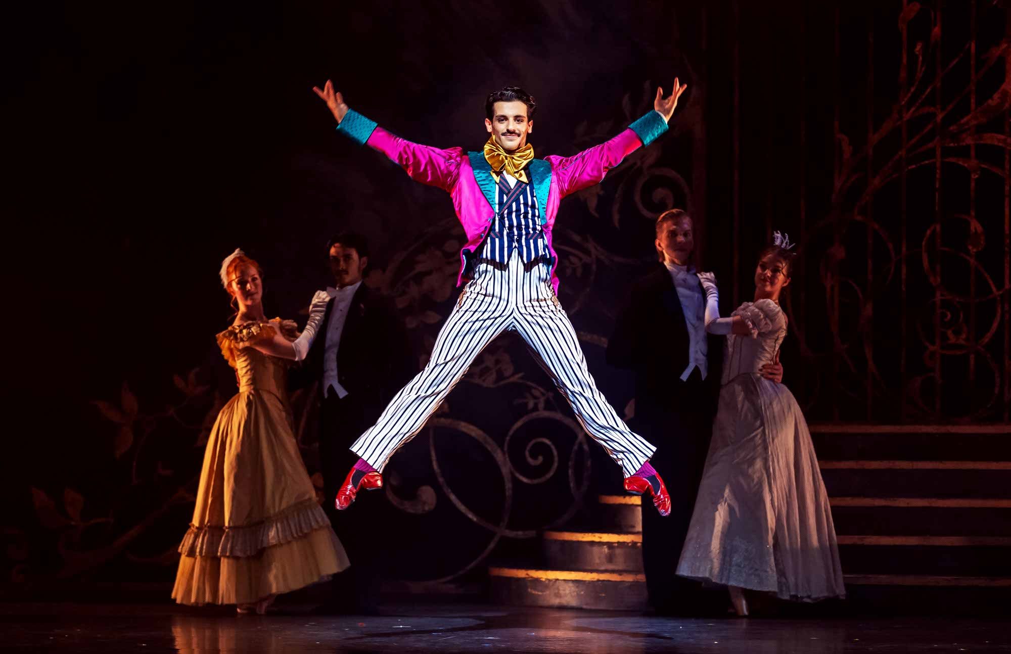A performer in a vibrant pink jacket and striped pants leaps mid-air on stage, arms outstretched, while other dancers in elegant period costumes stand behind him against a dark, theatrical backdrop.